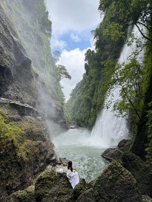 Standing on rocks at the foot of a tall waterfall in a green gorge in Meghalaya